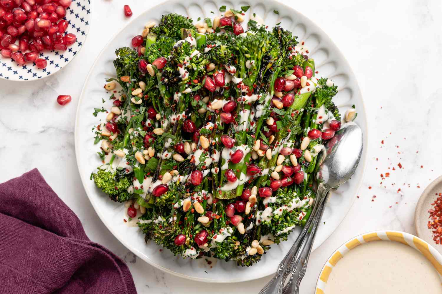 Grilled Broccolini with Lemon Tahini Dressing, Pomegranate Arils, and Pine Nuts on a Plate with Serving Spoon, Surrounded by Bowl of Pomegranate and Bowl of Tahini