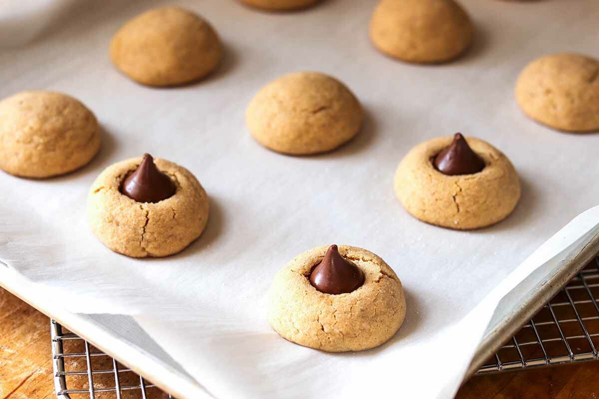 Baked peanut butter blossom cookies on a cookie sheet with hershey's kisses being placed on top.