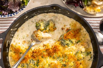 Skillet chicken divan in a cast iron skillet (with some already served) at a table setting with a bowl of salad, a bowl of parmesan, a small bowl of chopped parsley, and a utensils