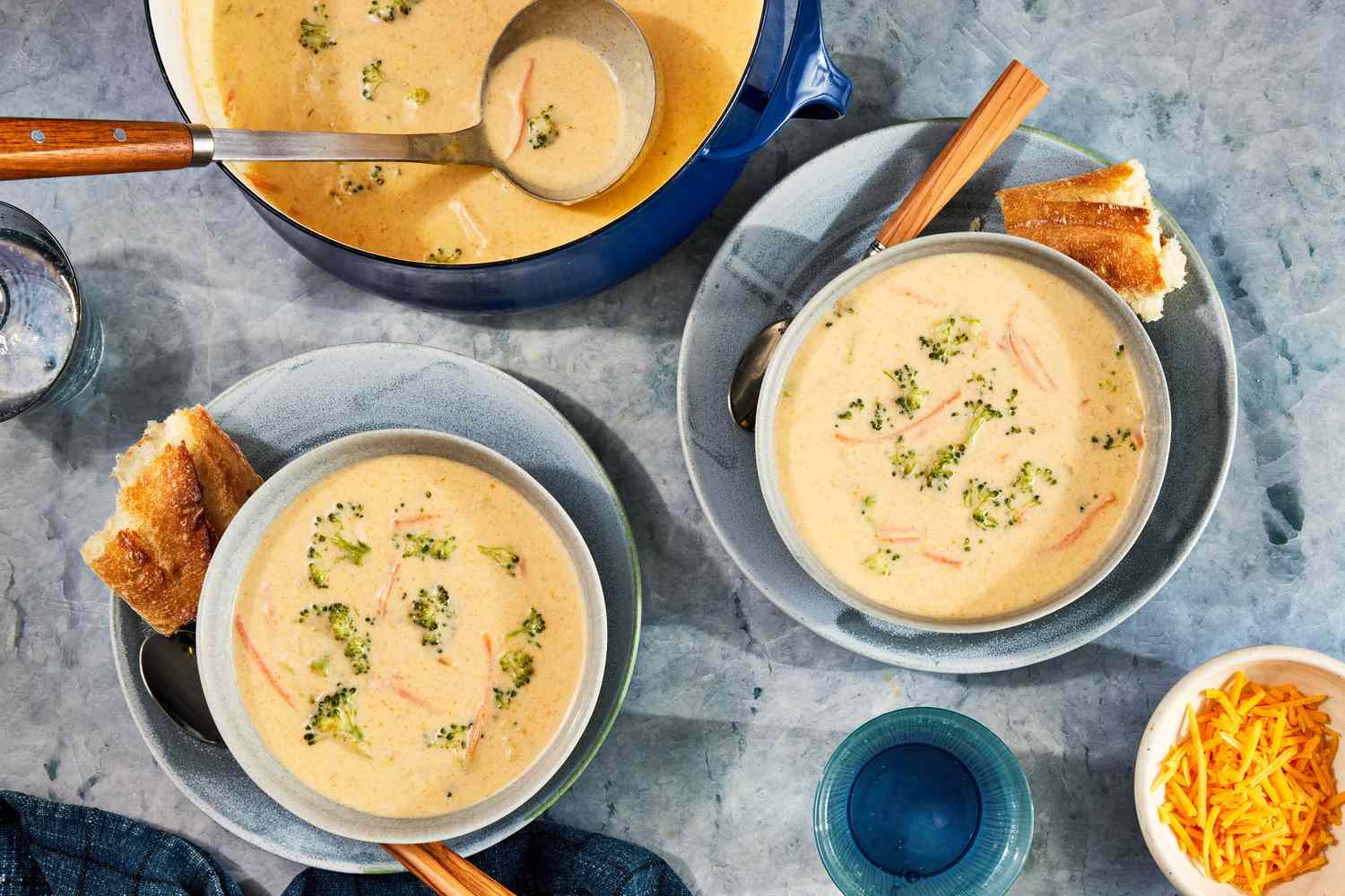 Two bowls of copycat Panera broccoli cheddar soup, each on a plate with a piece of baguette and a spoon, and in the surroundings, a pot with more soup, glasses of water, a blue kitchen towel, and a small bowl of shredded cheese