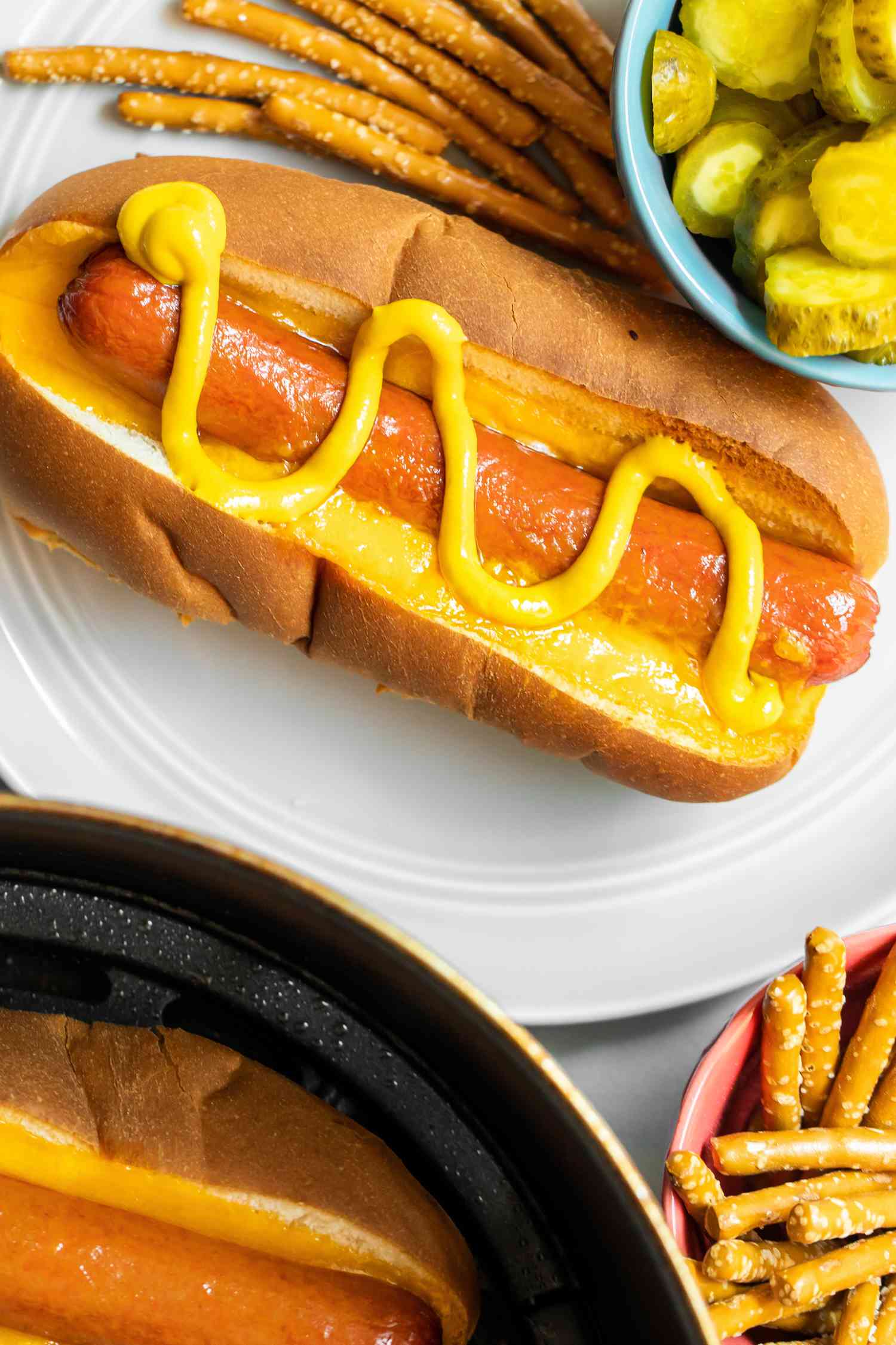Air Fryer Hot Dog in a Bun on a Plate with Mustard Drizzled on Top and a Side of Pickles and Pretzels