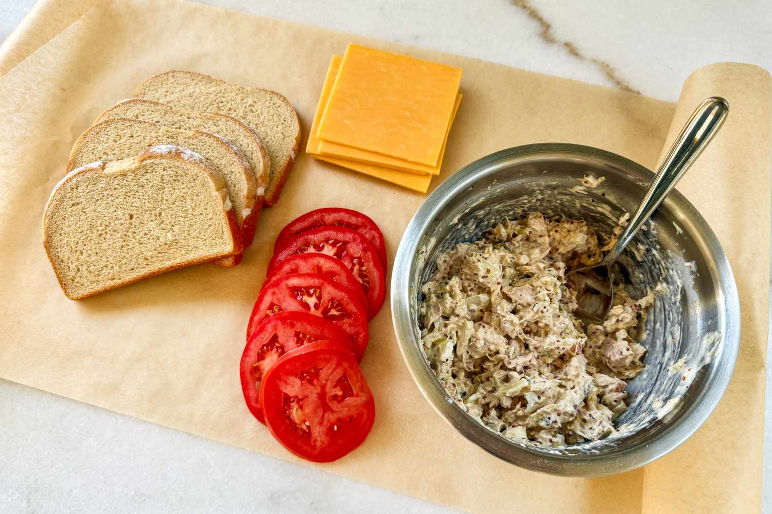 Tuna melt components arranged on a piece of parchment paper: the tuna salad, tomato slices, cheddar slices, and bread
