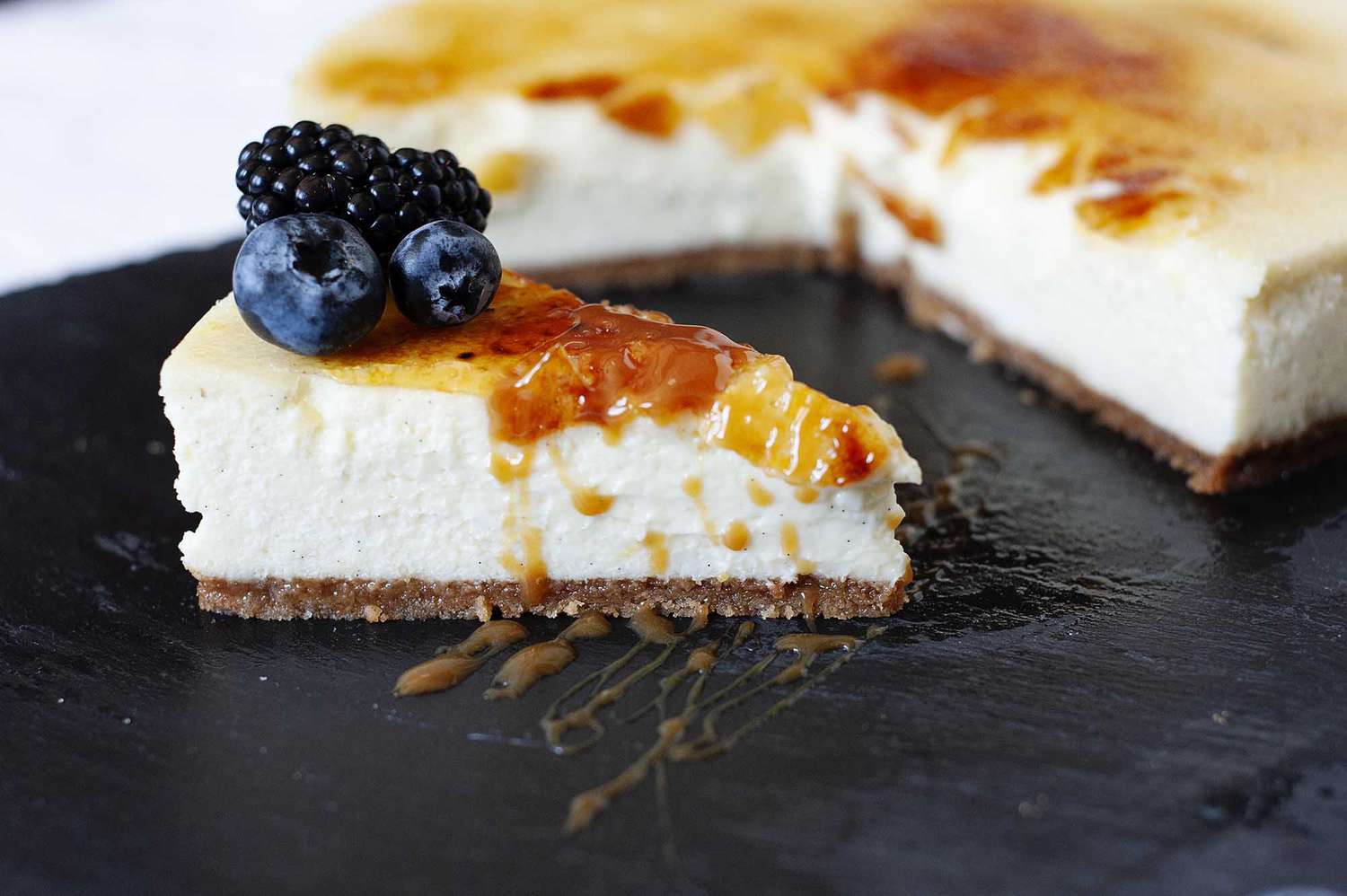 Vanilla Bean Cheesecake with brittle sugar topping on a slate board wtih a slice cut out and fruit nearby.