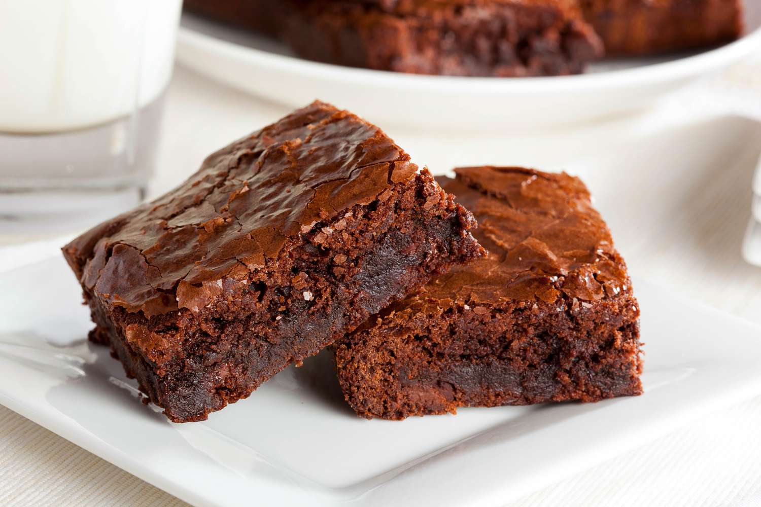 Two brownies on a square plate, and in the background, a glass of milk and a plate with more brownies