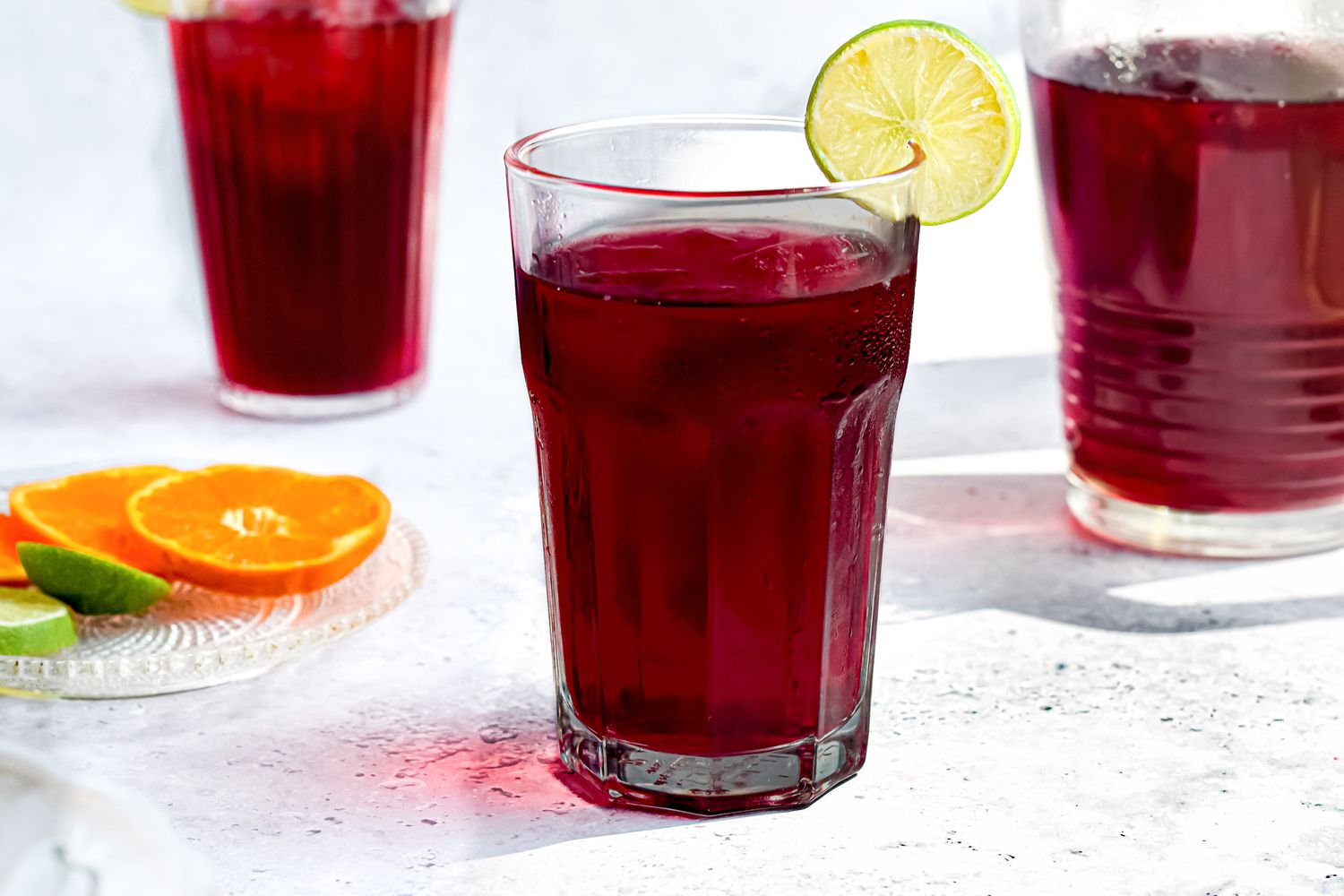 Agua de Jamaica (Hibiscus Tea) Served with Ice and a Lime Slice Next to a Small Plate of Sliced Oranges and Limes, a Bowl with Dried Jamaica Flowers, and a Pitcher 