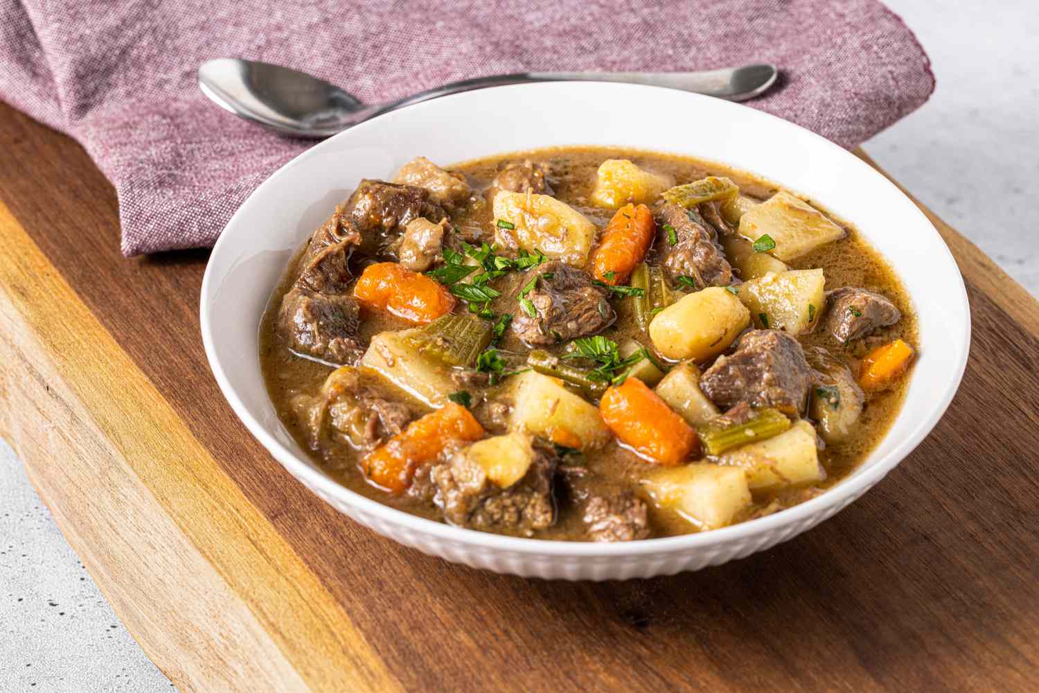Bowl of Easy Beef Stew Toped with Some Parsley and in the Background, a Kitchen Towel with a Spoon on Top
