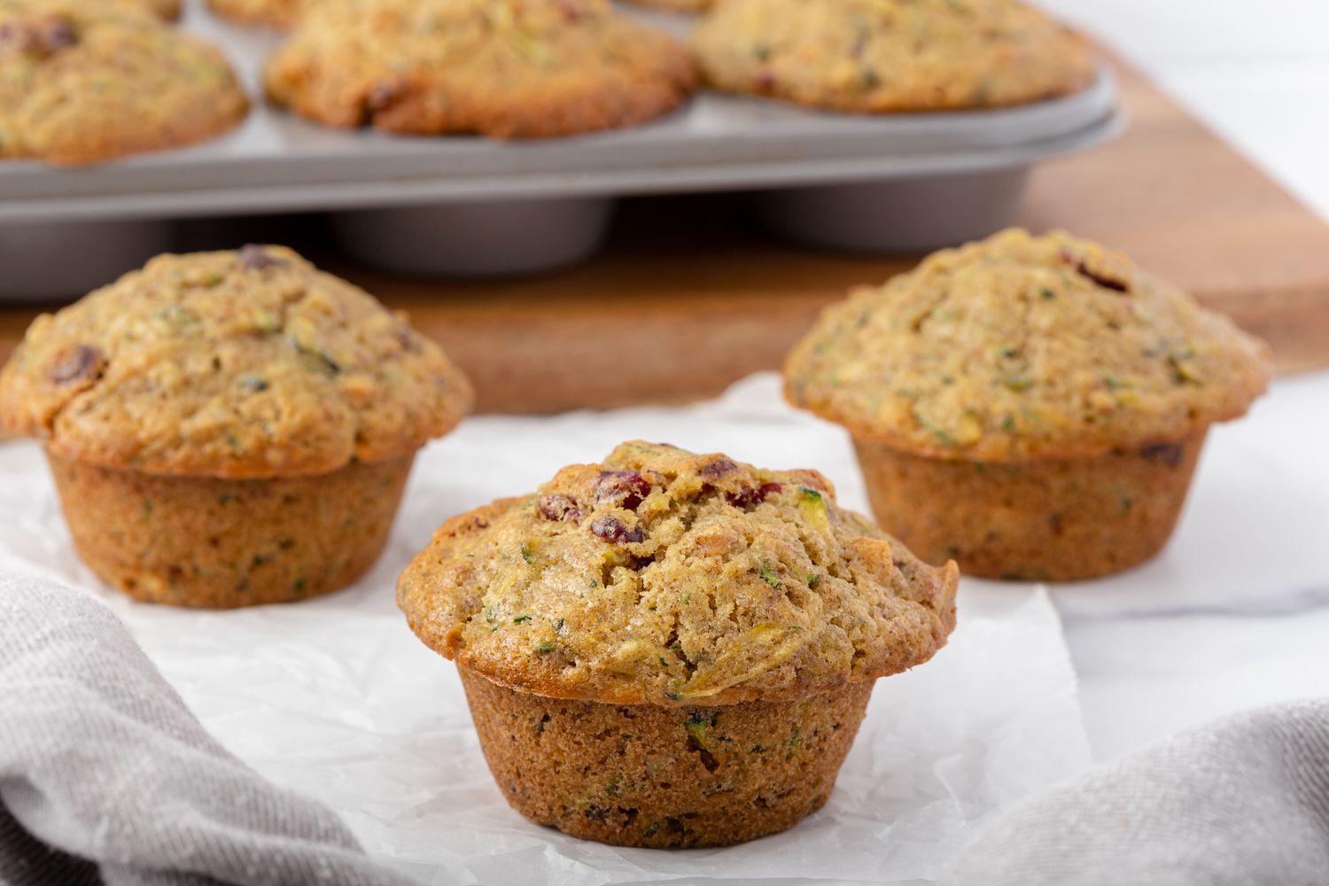 Three zucchini bread muffins set in front of a muffin tin with muffins inside.