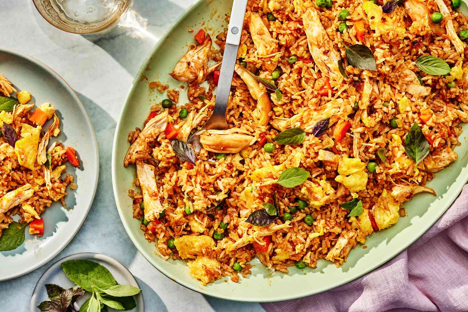 Thai fried rice on a platter at a table setting with a serving of fried rice on a plate, a small plate with Thai basil, a glass of water, and a table napkin