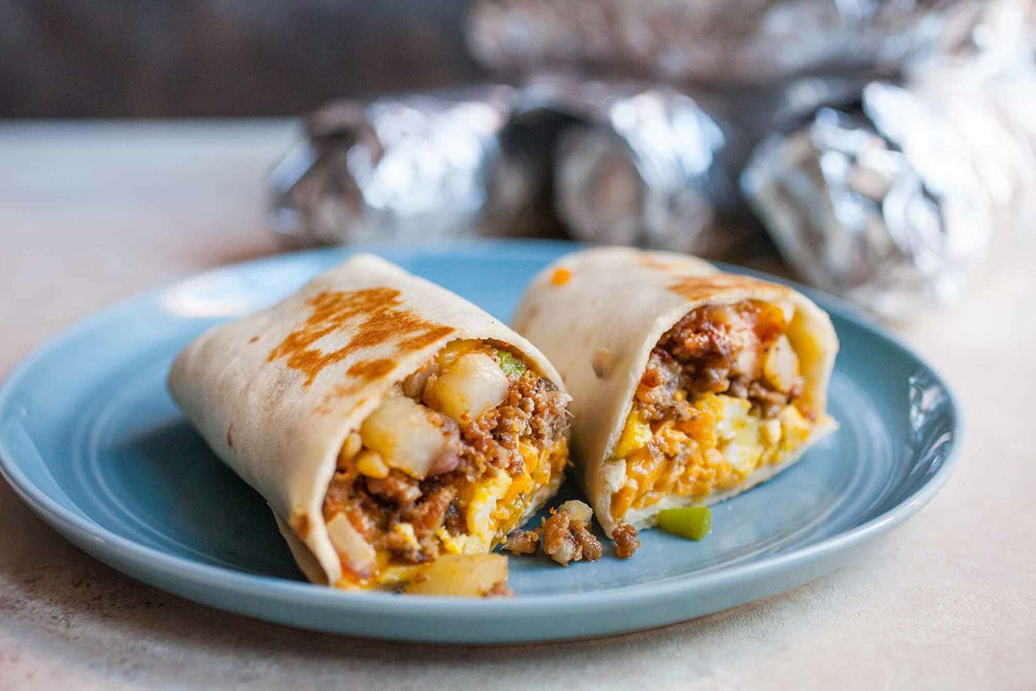 A breakfast burrito, cut in half in the middle, on a plate with foil-wrapped breakfast burritos in the background