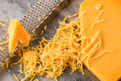 Overhead view of a large block of cheddar cheese, a shredder and pile of shredded cheese on a marble countertop