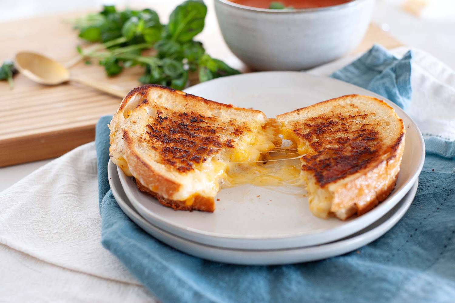The perfect grilled cheese sandwich cut in half and split open on a plate with a blue napkin underneath it and a bowl of tomato soup in the background.