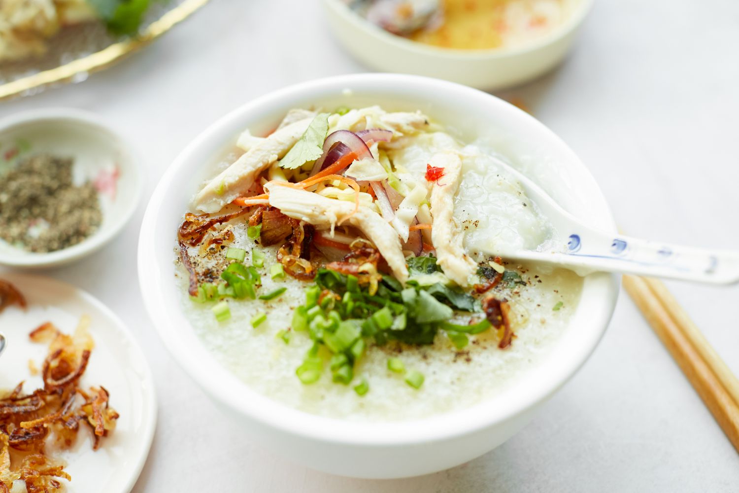 Chào Gá (Vietnamese Rice Porridge) in a Bowl with Soup Spoon, Surrounded by Bowl of Seasoning and Small Plate of Fried Onions