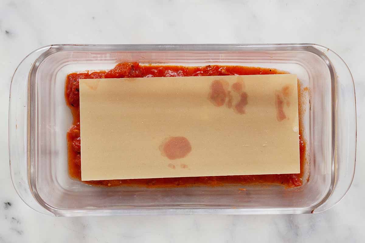 A lasagna noodle is atop chunky tomato sauce and sliced sausage in a glass loaf pan.