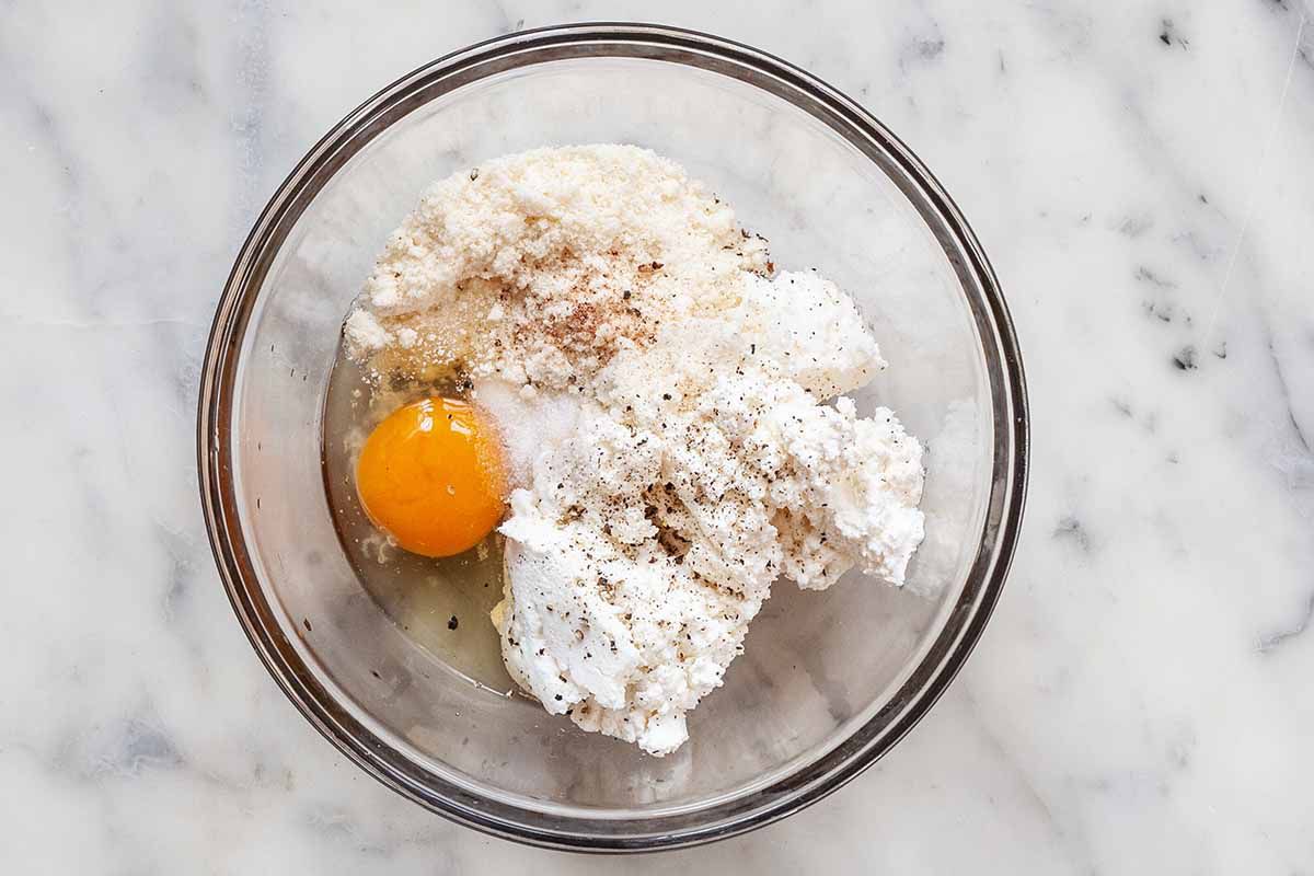 Small glass bowl with ricotta, parmesan, spices and an egg inside.