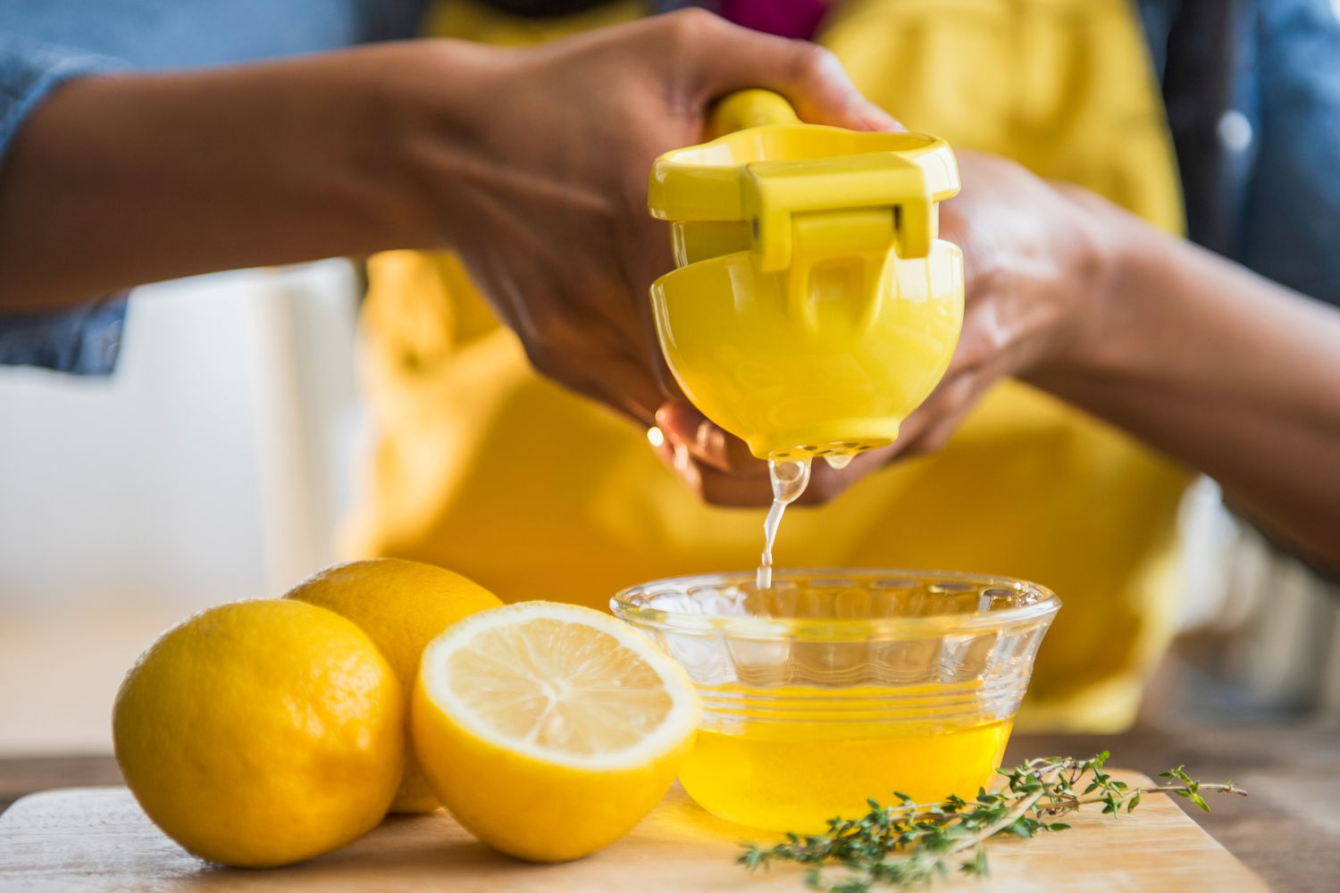 A lemon being juiced with a hand juicer. The juice is going into a small glass bowl on a cutting board with more lemons and sprigs of thyme