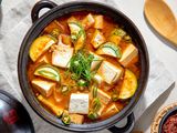 Overhead View of Doenjang Jjigae in a Clay Pot on a White Kitchen Towel and Surrounded by a Bowl of Rice, a Small Bowl of Sauce, and a Wooden Spoon
