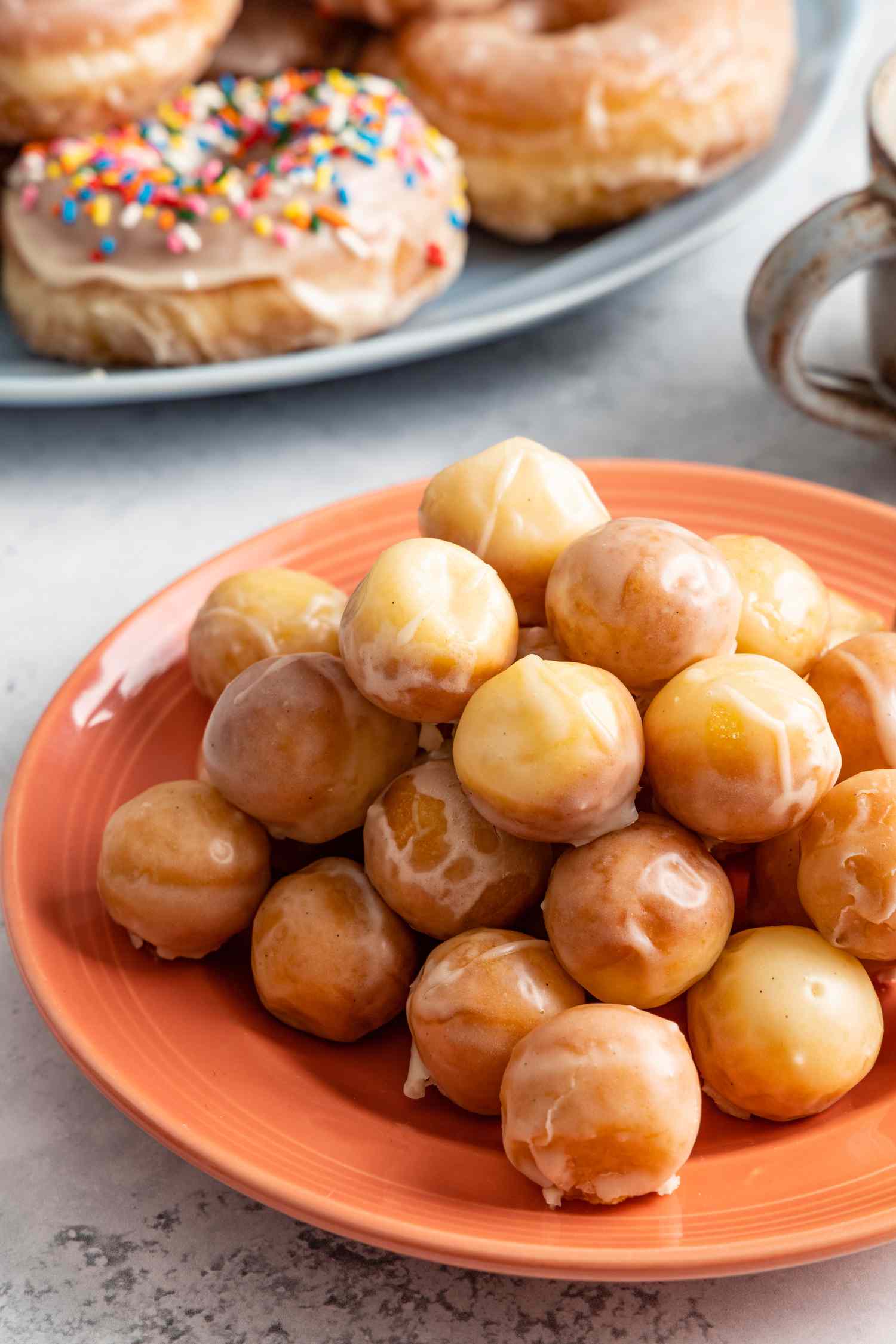 Platter of Glazed Donut Holes, and in the Background, a Platter of Glazed Donuts and a Mug of Coffee