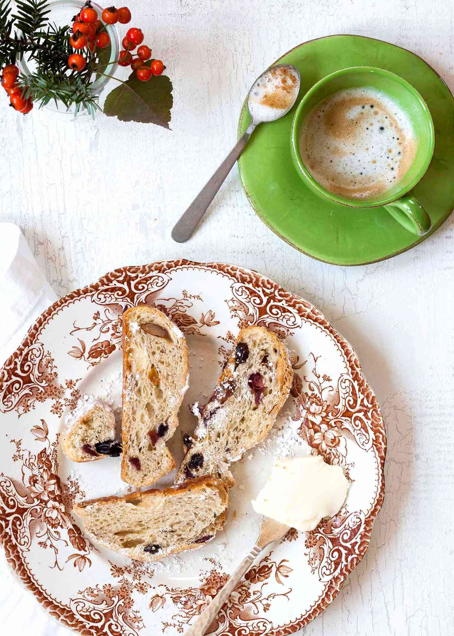 Stollen Christmas Bread filled with rum-soaked dried fruit coated with powdered sugar and sliced on a platter with a fine mesh sieve with powdered sugar and a cup of coffee in a green mug.