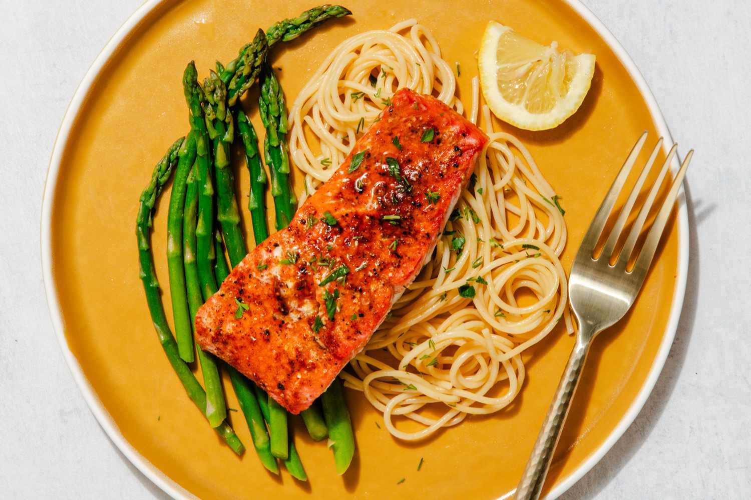 A vibrant fillet of roasted salmon served over asparagus and plain spaghetti noodles, all on a bright yellow plate