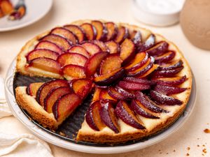 german plum tart (missing a slice) on a plate at a table setting with a kitchen linen, a slice of tart on a plate with a fork, and a bowl of sugar