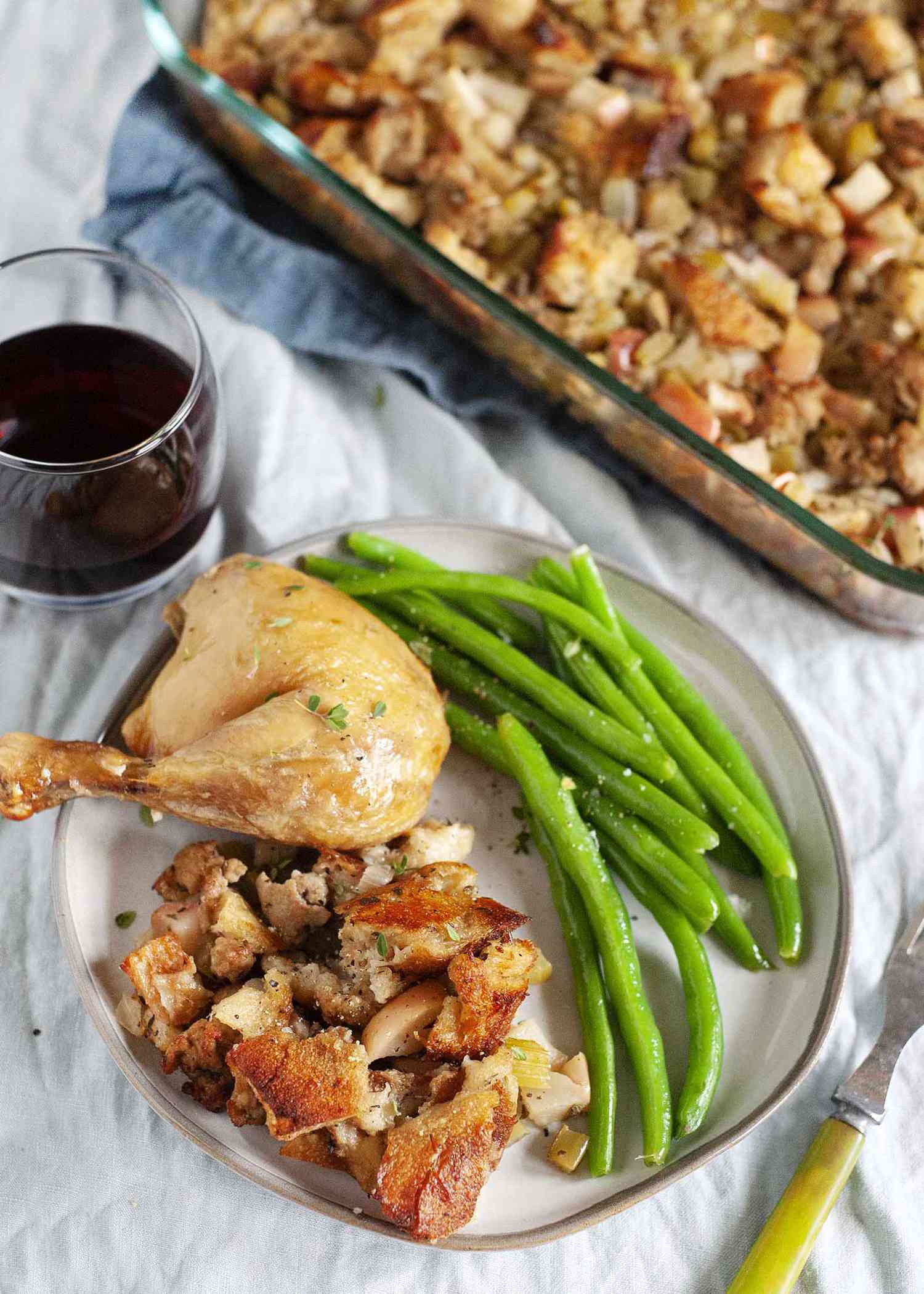 A baking dish with classic sage, sausage and apple stuffing next to plate of stuffing, chicken leg, and green beans. There is a glass of red wine nearby.