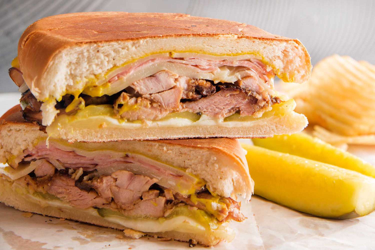 A side view of a cuban sandwich with pickles and chips in the background