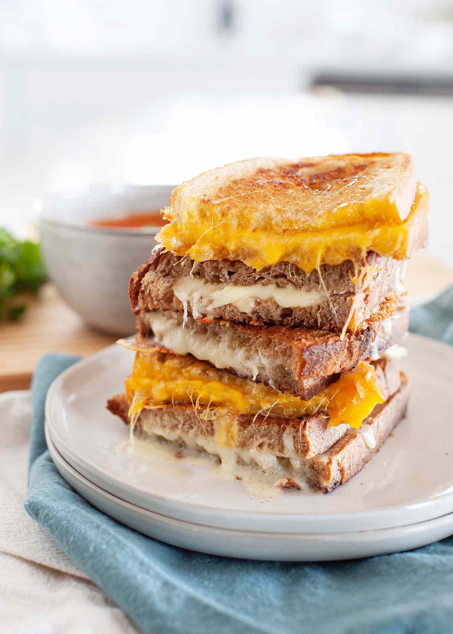 Grilled cheese sandwiches cut in half and stacked ontop of each other. Alternating with orange and white cheese with a bowl of tomato soup in the background.
