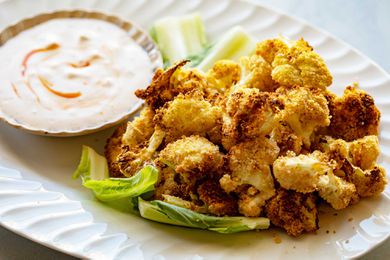 Platter of golden air fried crispy cauliflower heaped on the right of the platter. A small bowl of creamy dip swirled with hot sauce is placed next to the cauliflower on the platter. The cauliflower is resting on a few leaves of lettuce.