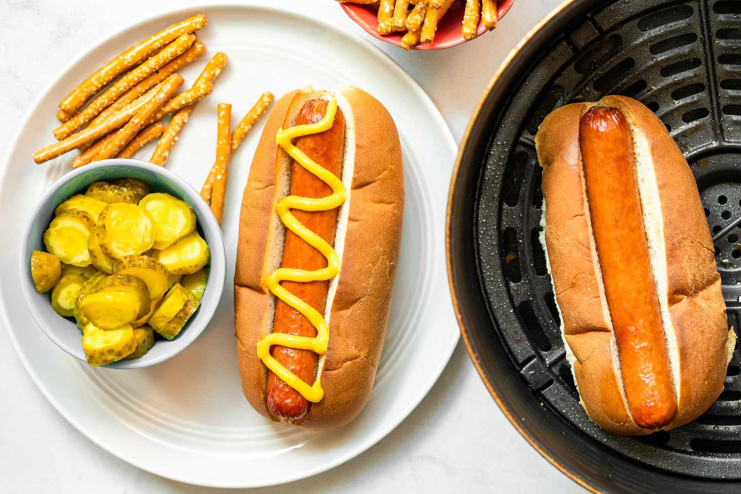A Plate of Air Fryer Hot Dog in a Bun Topped with Mustard and a Side of Pickles and Pretzels. Next to It Is the Air Fryer Basin with Another Hot Dog in a Bun and a Bowl of Pretzels.