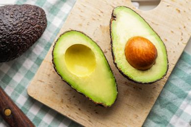 A perfectly ripe avocado cut in half, showing the pit inside. The avocado is on a small cutting board, which is sitting on a green checked kitchen towel