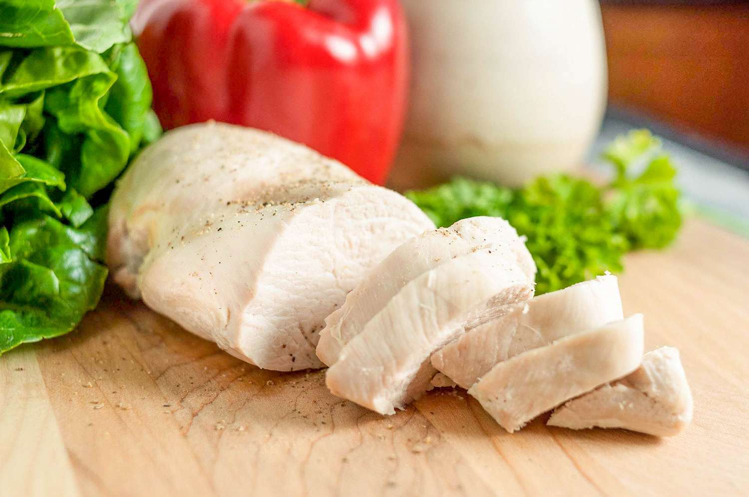 A cooked boneless skinless chicken breast on a cutting board, partially sliced with a bell pepper in the background