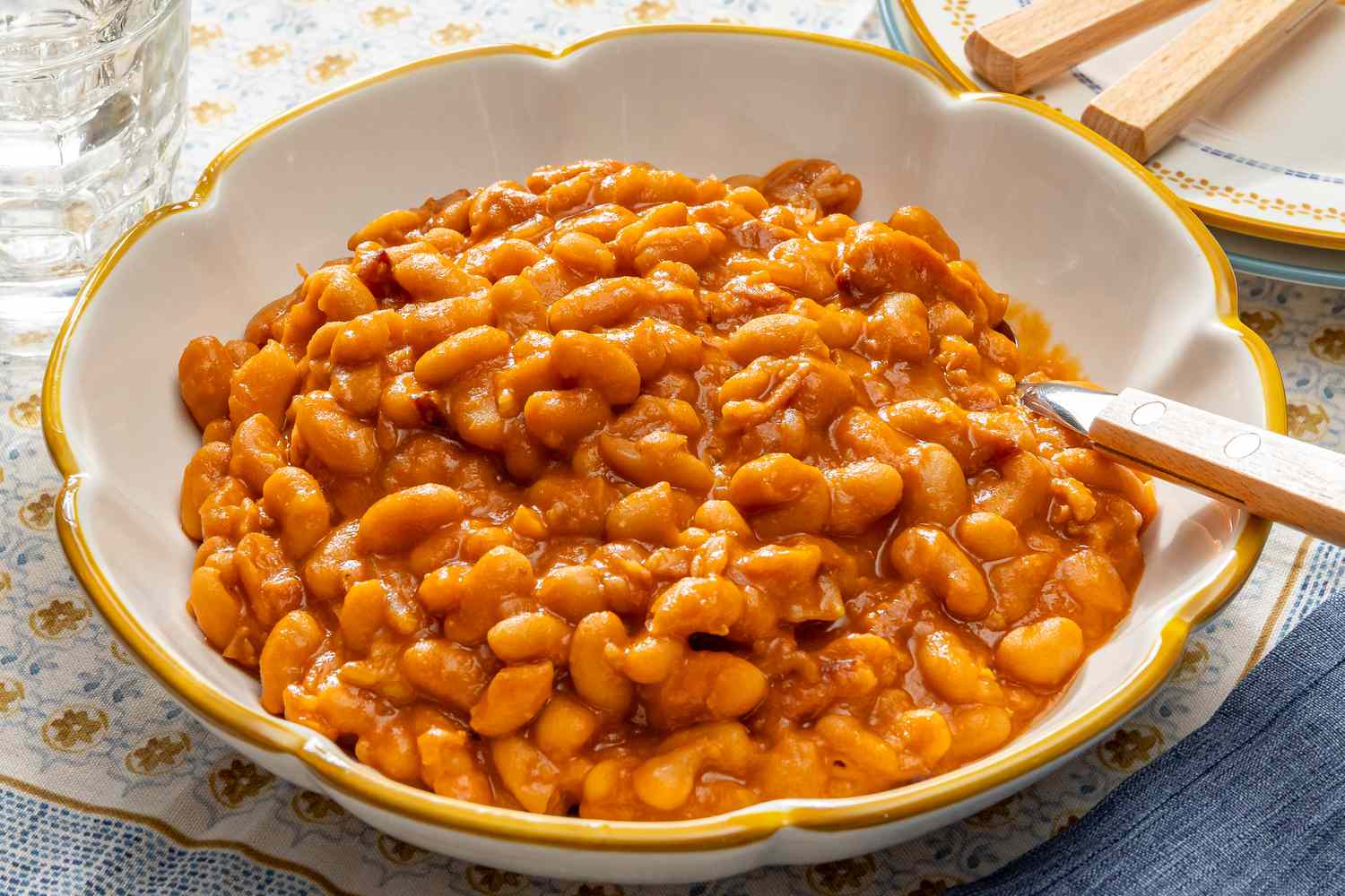 A bowl of baked beans with sauce and a spoon on a patterned tablecloth