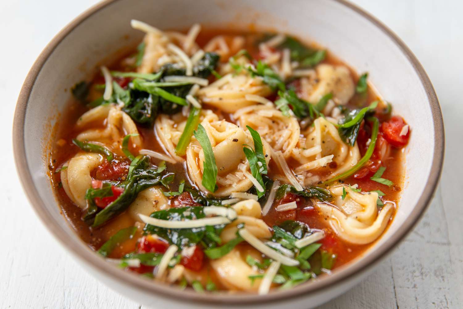 A serving of tortellini soup with a tomato based broth in a bowl, topped with parmesan cheese