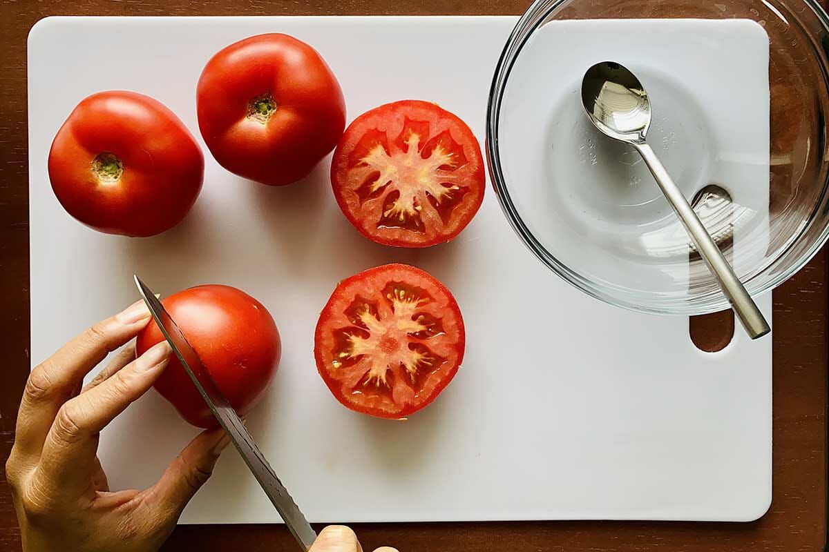 Tomatoes being halved on a cutting board with a glass bowl and spoon set to the right.