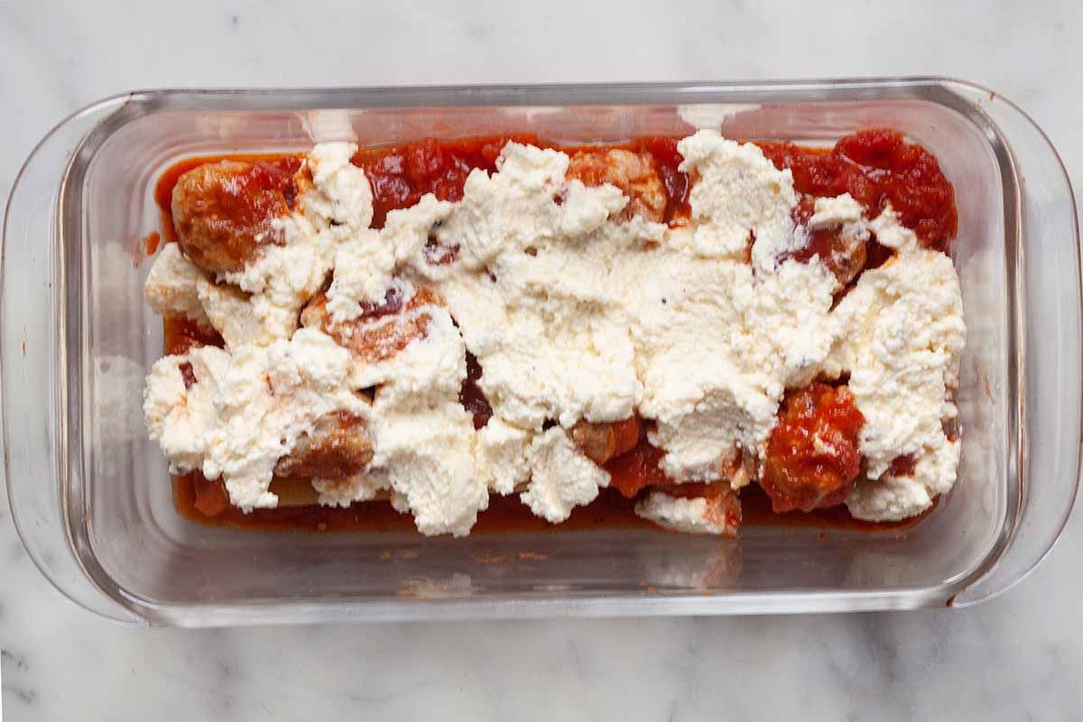 A glass loaf pan with ricotta cheese dotted on top sliced sausage and tomato sauce in a glass loaf pan for small batch lasagna.
