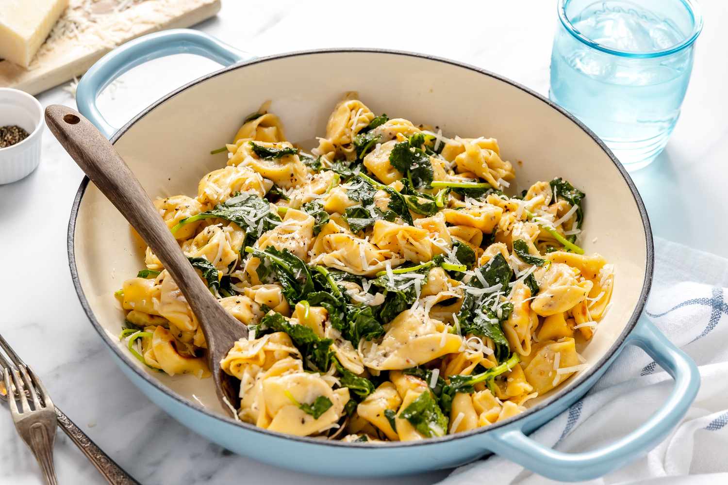 Skillet Cacio e Pepe Tortellini With Wilted Greens in a Pot With a Serving Spoon, All on a White Kitchen Towel on the Counter, and in the Surroundings, a Glass of Water, a Block of Pecorino Romani Cheese on a Cheese Board, a Small Bowl of Cracked Pepper, and Two Forks on the Counter