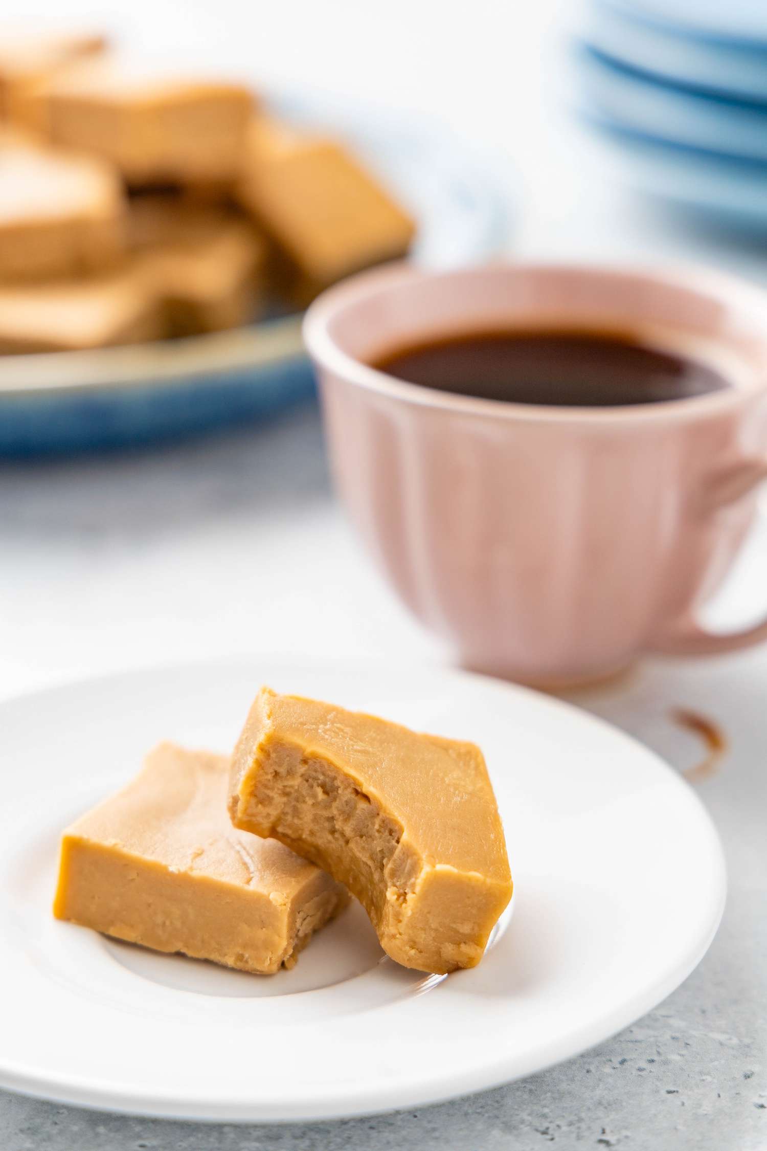 Serving of penuche on a small plate (one piece missing a bite), and in the background, a teacup with coffee and a plate with more penuche