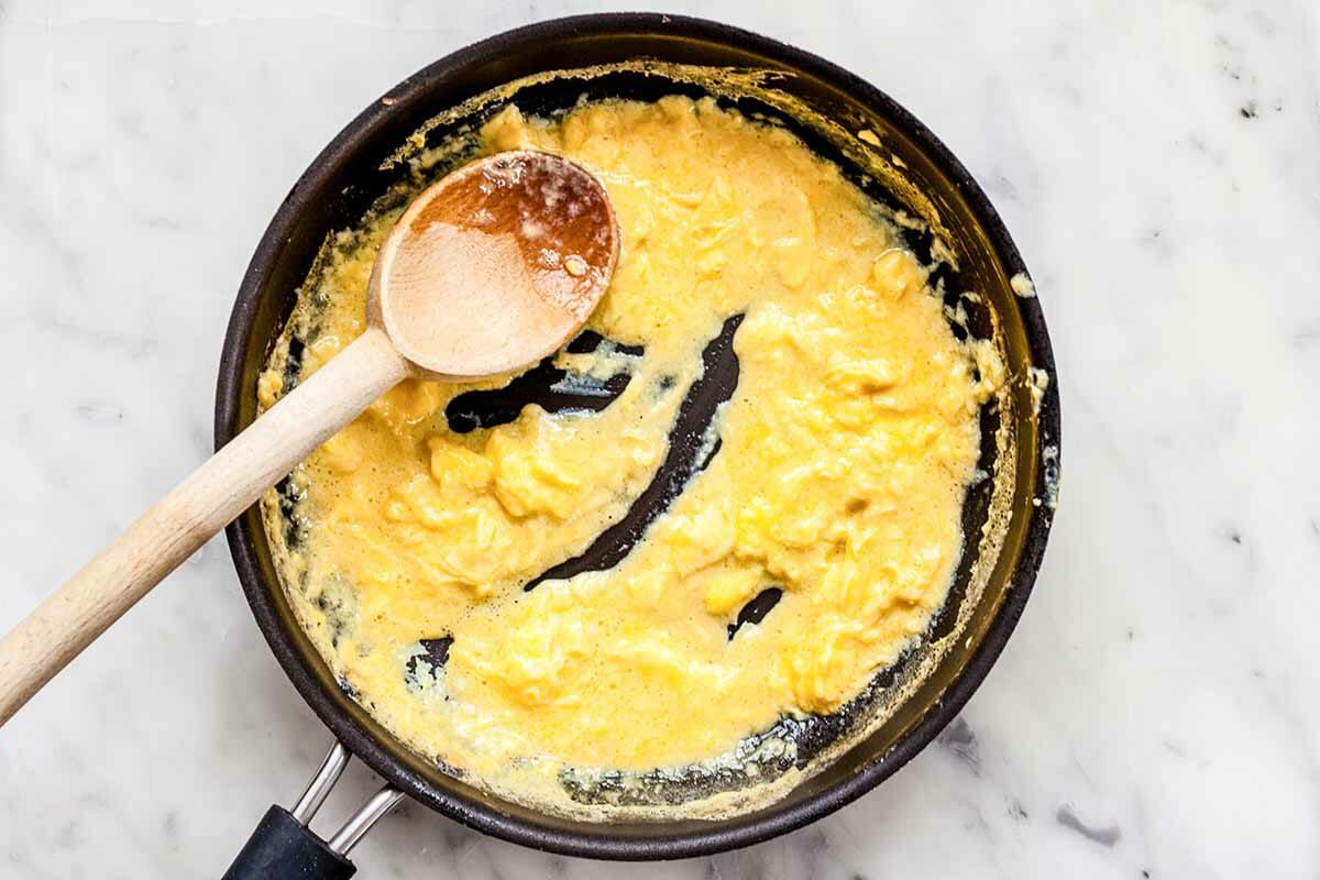 Scrambled eggs cooking on a skillet and being stirred with a wooden spoon