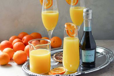 Prosecco, orange juice, orange slices and three glasses of the best mimosas are on a tray. Whole oranges are to the left of the tray.