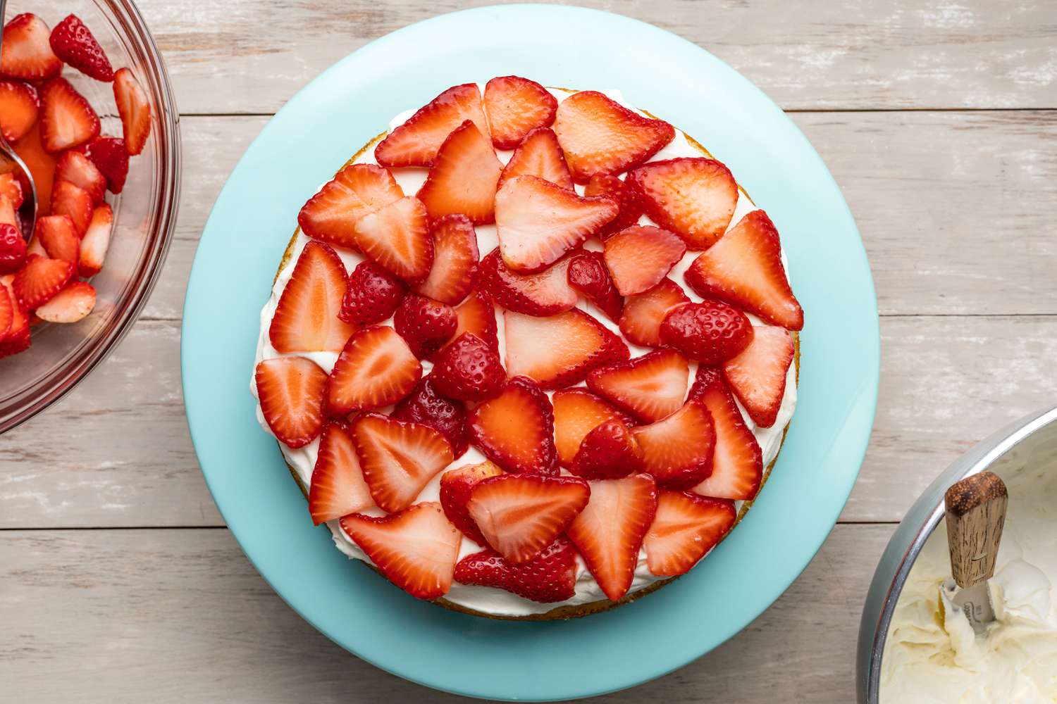 Strawberries Added on Top of Whipped Cream (Cake on Cake Stand Next to a Bowl of Sliced Strawberries and the Bowl of Whipped Cream Frosting)