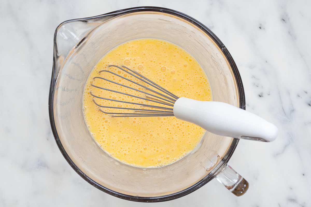 A large glass liquid measuring cup is on a marble countertop and a whisk with a white handle is inside. A smooth egg filling for the best deep-dish bacon and cheddar quiche