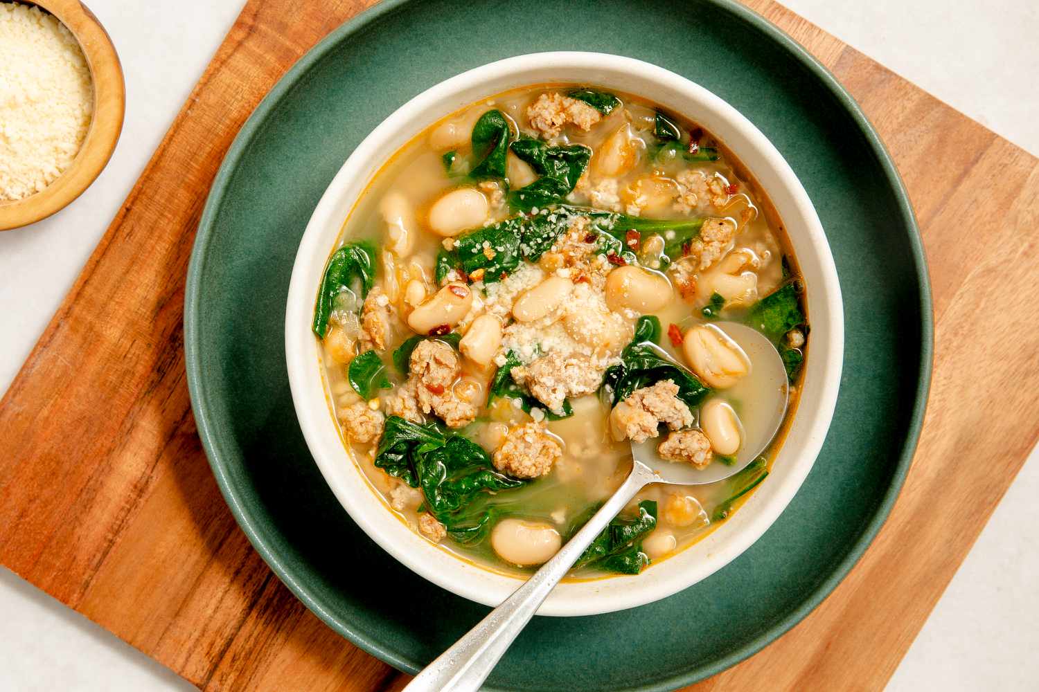serving of 15-Minute Sausage White Bean Soup in a white bowl on a dark green plate on a wooden serving board next to a bowl of shredded parmesan