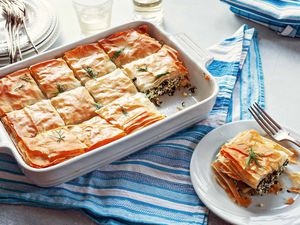 Slice of Greek Spinach Pie on a white plate, with a fork, a glass of white wine, and the casserole dish in the background,