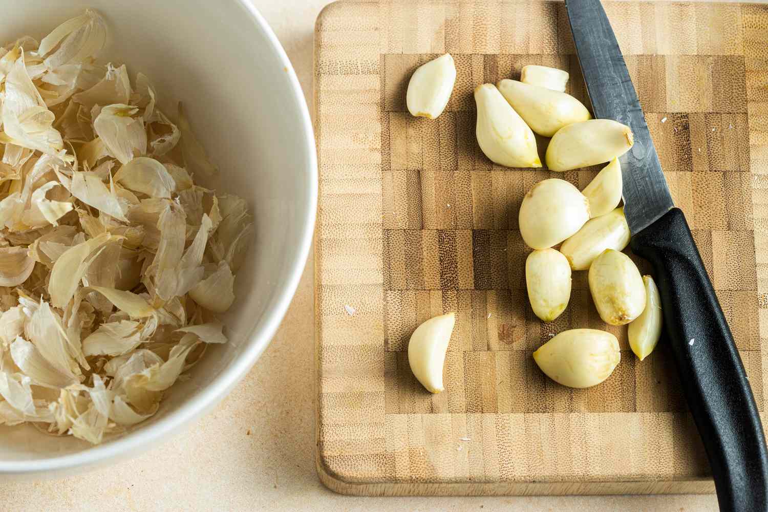 Peeled Garlic on Cutting Board with Knife and Peels in Bowl