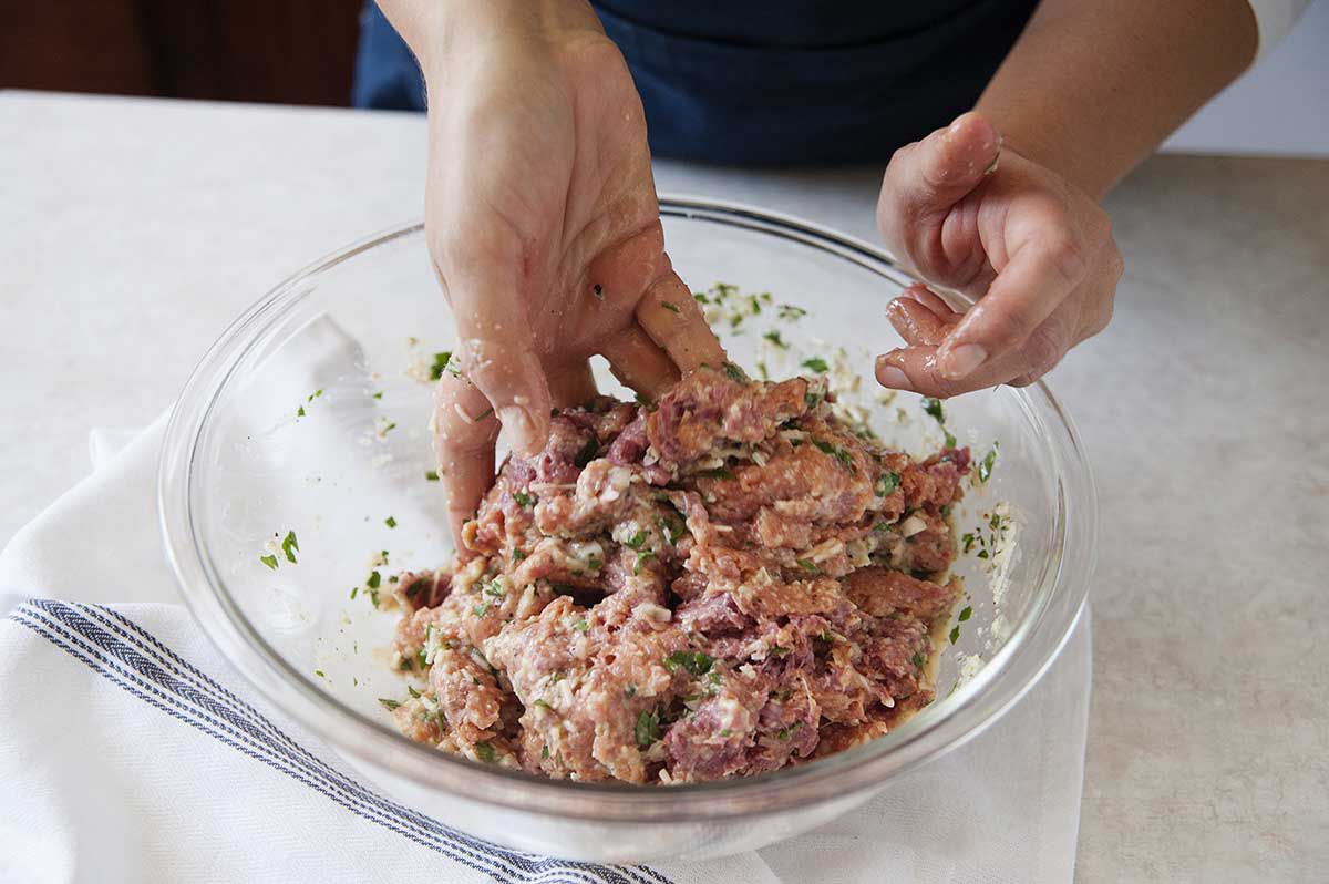 Eggs, breadcrumbs, herbs and cheese in a bowl being stirred together with two different kinds of meat to make slow cooker meatballs.