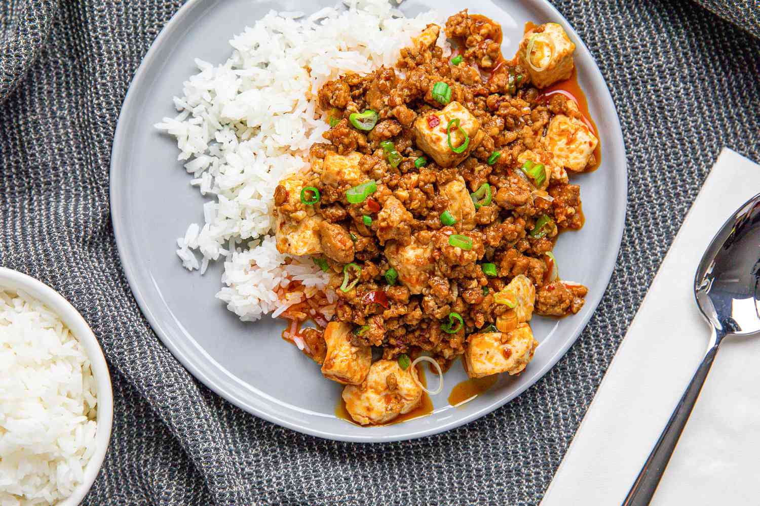 Plate of Mapo Tofu Served Over Rice at a Table Setting With a Spoon on a Napkin and a Bowl of Rice 