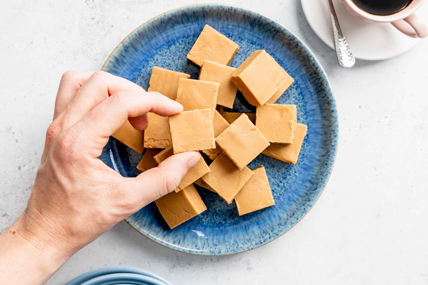 Someone lifting a piece of penuche from a plate next to a teacup with coffee 