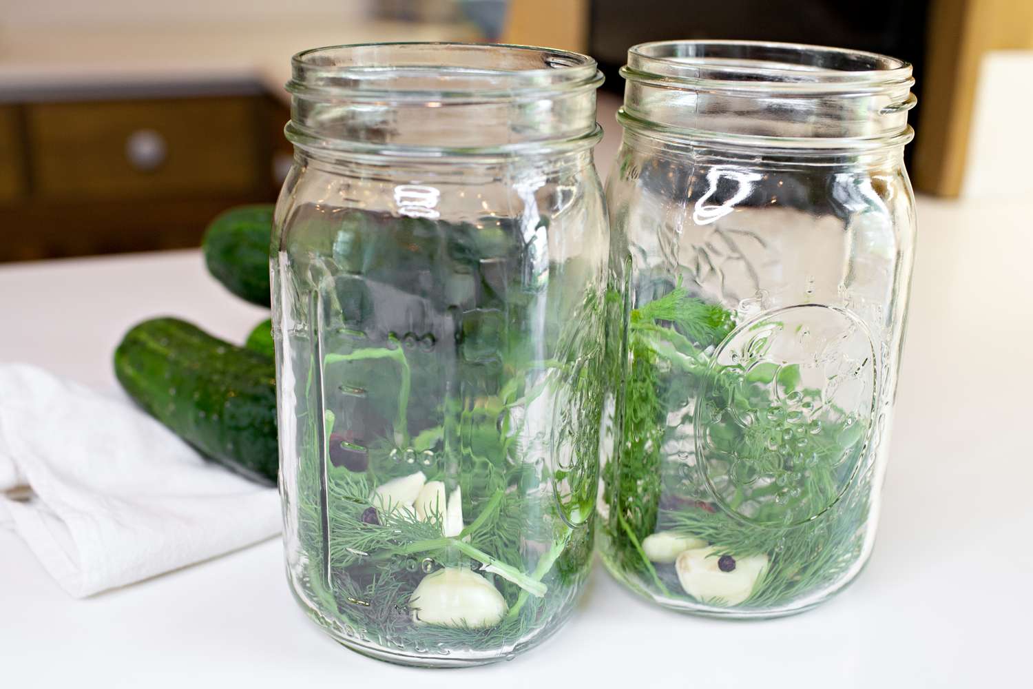 Garlic Cloves, Black Peppercorn, and Fresh Dill Added to Mason Jars. In the Background, Cucumbers on the Counter for Pickles Recipe