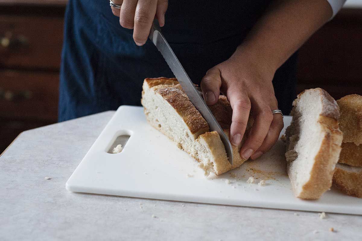 Woman slicing sourdough bread on a baking sheet to make thanksgiving sausage stuffing.