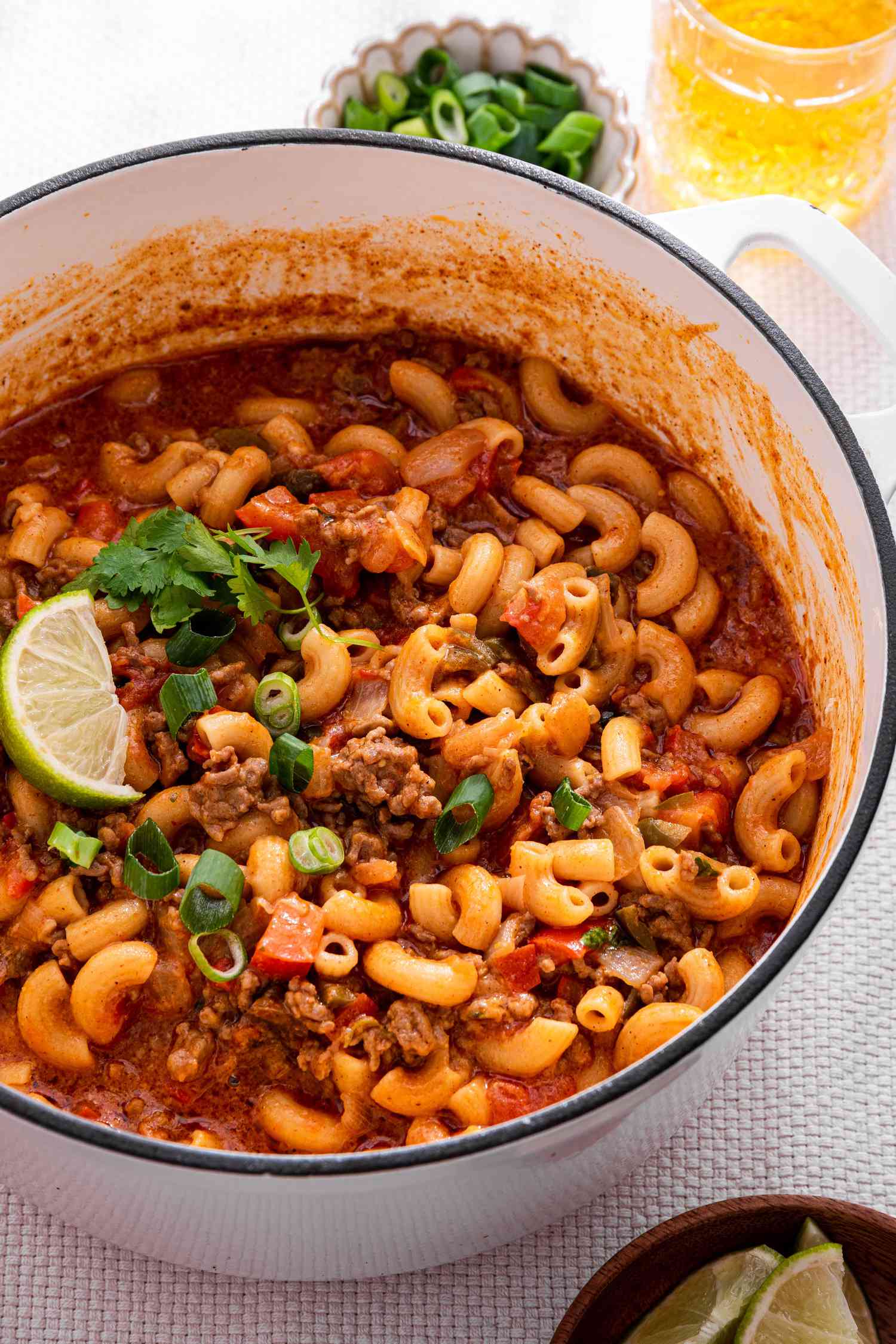One-Pot Taco Pasta in a Pot, and in the Surroundings, a Bowl With Lime Wedges, a Bowl With Sliced Green Onions, a Bowl With Sour Cream, Glasses With a Drink, and a Mustard Colored Table Napkin